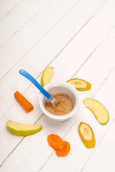 Vista de alto ângulo de alimentos para bebês na tigela com colher e frutas fatiadas e cenoura no fundo de madeira branca — Fotografia de Stock