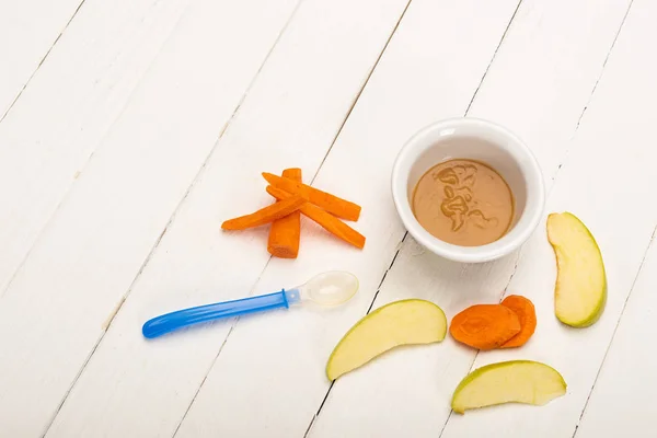 High angle view of baby food in bowl near spoon and sliced ingredients on white wooden surface — Stock Photo