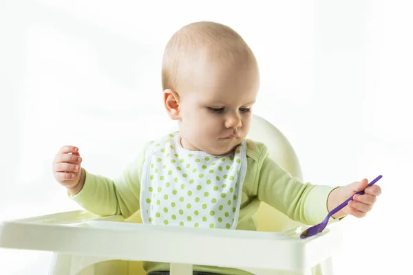 Cute baby holding spoon while sitting on feeding chair on white background — Stock Photo