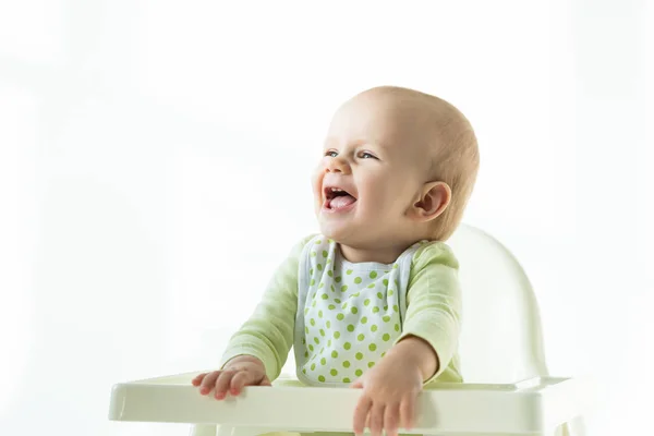 Веселый ребенок в нагруднике, смотрящий на стул для кормления на белом фоне — стоковое фото