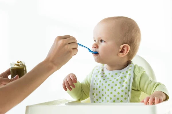 Мать с банкой детского питания и ложкой кормления младенца на кормящем стуле на белом фоне — стоковое фото