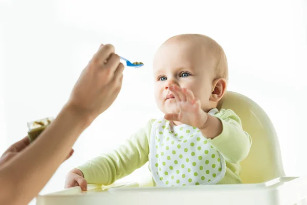 Selective focus of cute baby pulling hand to mother with spoon of baby nutrition isolated on white — Stock Photo