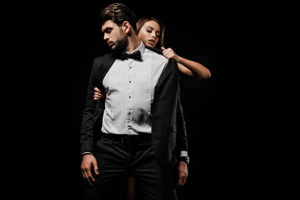 Mujer apasionada quitándose la chaqueta de hombre guapo en traje aislado en negro - foto de stock