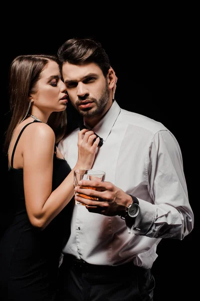 Sexy woman touching bow tie of handsome man with glass of whiskey isolated on black — Stock Photo