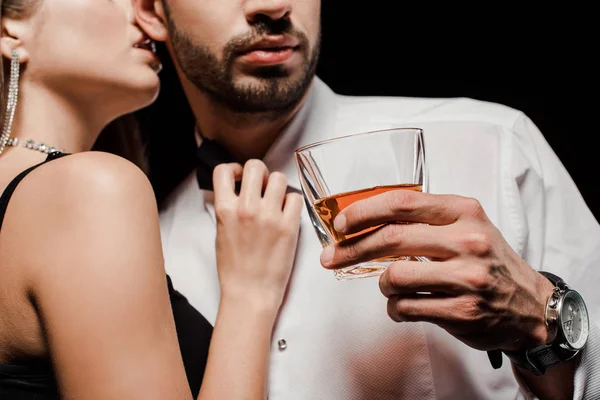 Cropped view of woman touching bow tie and kissing man with glass of whiskey isolated on black — Stock Photo