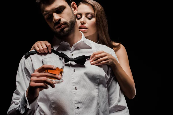 Attraktive Frau trägt Fliege auf Mann mit Glas Whiskey isoliert auf schwarz — Stockfoto