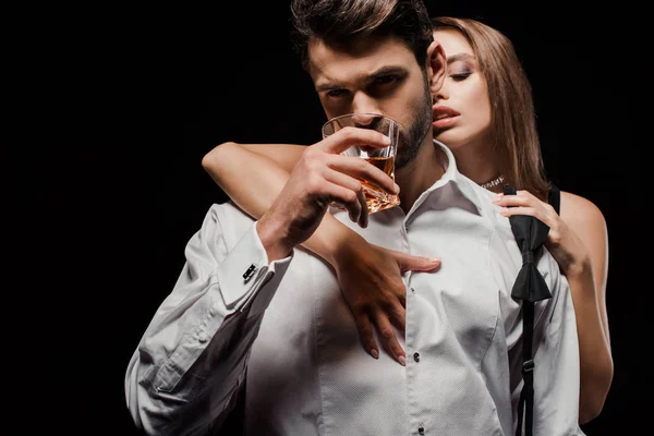 Attractive woman holding bow tie and touching man drinking whiskey isolated on black — Stock Photo