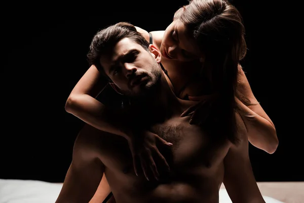 Sinnliche Frau berührt hemdlosen Mann auf Bett isoliert auf schwarz — Stockfoto