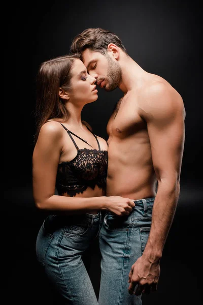 Вид сбоку мускулистого мужчины, целующегося с молодой женщиной в кружевном бюстгальтере на черном — стоковое фото