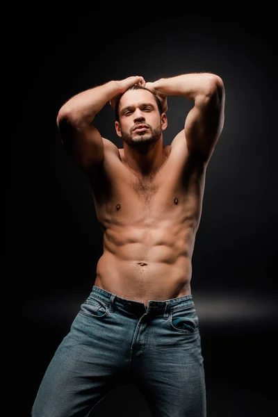 Handsome and muscular man in jeans touching hair on black — Stock Photo