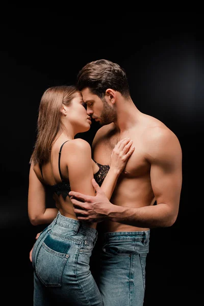 Вид збоку без сорочки чоловік із закритими очима цілує привабливу жінку в джинсах на чорному — стокове фото