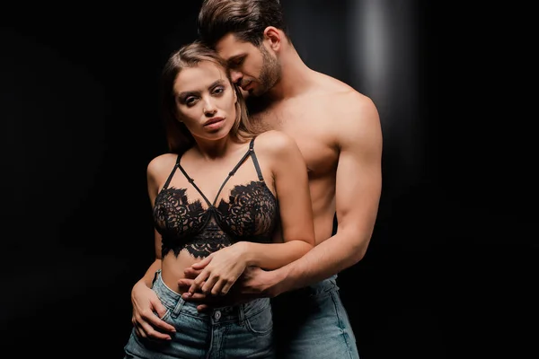 Shirtless man hugging attractive woman in jeans and bra on black — Stock Photo