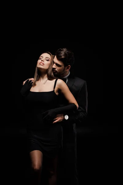 Hombre guapo en traje abrazando atractiva mujer aislado en negro - foto de stock