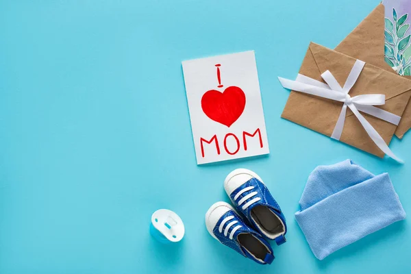 Top view of i love mom lettering on greeting card near baby hat, pacifier and booties on blue background — Stock Photo