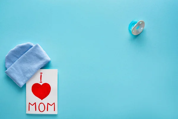 Visão superior de eu amo a mãe lettering no cartão de saudação perto de chapéu e chupeta no fundo azul — Fotografia de Stock