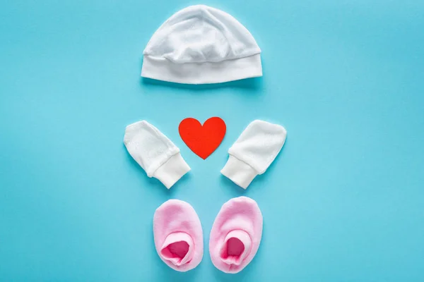 Vista superior de manoplas de bebé, sombrero y botines con corazón de papel sobre fondo azul, concepto de día de las madres - foto de stock