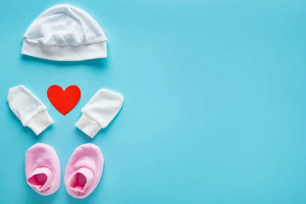 Draufsicht auf Baby-Fäustlinge, Booties und Hut mit herzförmigem Papier auf blauem Hintergrund, Muttertagskonzept — Stockfoto