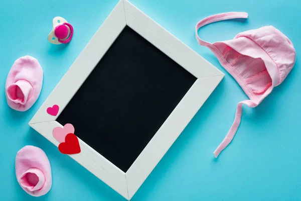 Top view of empty chalkboard with pink baby clothes and pacifier on blue background, concept of mothers day — Stock Photo