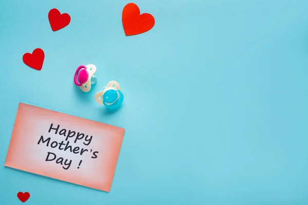 Vista superior de las madres felices letras del día en la tarjeta con chupetes y corazones de papel sobre fondo azul - foto de stock