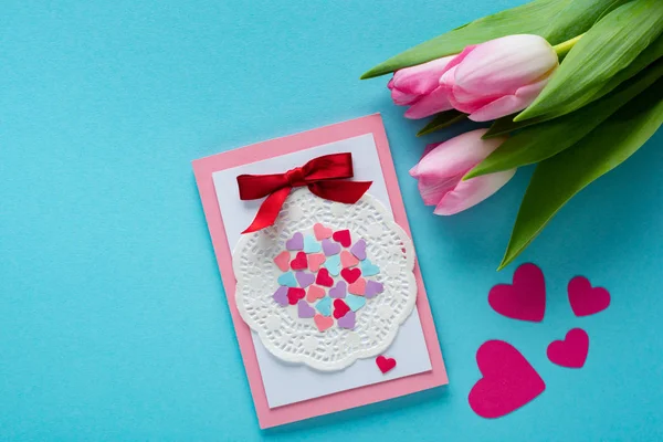 Top view of greeting card with tulips and paper hearts on blue surface — Stock Photo