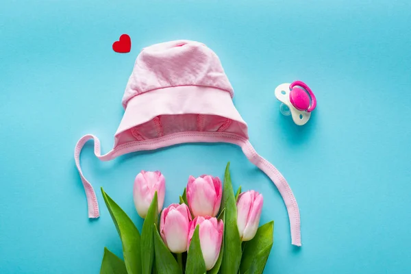 Top view of pink baby hat and tulips near pacifier on blue background, concept of mothers day — Stock Photo