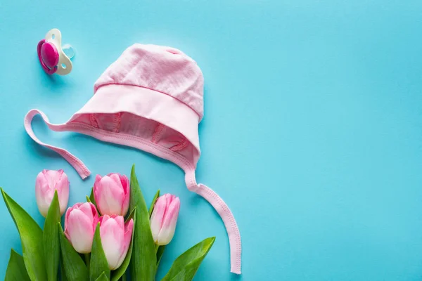 Top view of pink baby hat and pacifier near bouquet of tulips on blue background, concept of mothers day — Stock Photo
