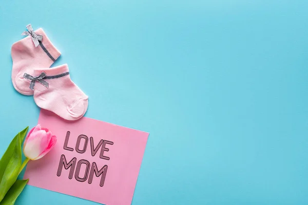 Vista superior de los calcetines rosados del bebé, tarjeta de felicitación con letras de la mamá del amor y tulipán en superficie azul - foto de stock