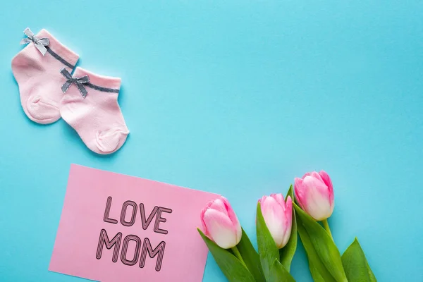 Vue du dessus des chaussettes roses pour bébés avec tulipes et lettrage de maman amoureuse sur la carte de vœux sur la surface bleue — Photo de stock