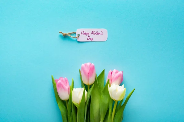 Vista dall'alto di etichetta di carta con felice giorno delle madri lettering e tulipani su sfondo blu — Foto stock