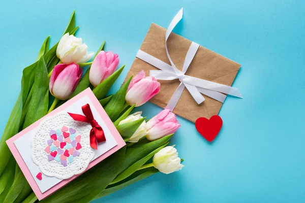 Top view of greeting card on tulips with envelope and heart shaped paper on blue background — Stock Photo