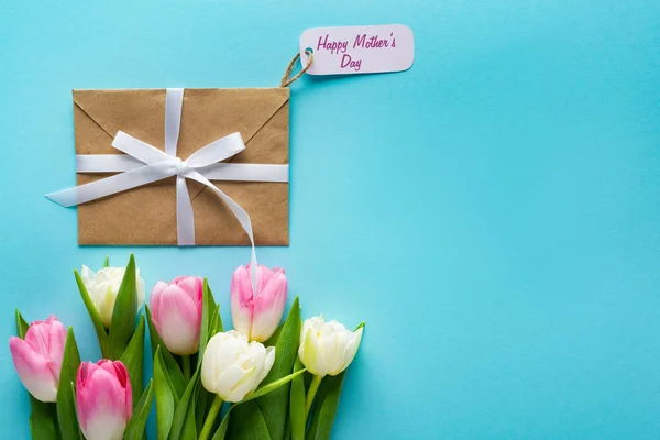 Top view of envelope with happy mothers day lettering on paper label and tulips on blue background — Stock Photo