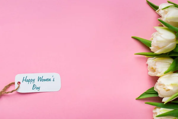 Vista superior de la etiqueta de papel con letras felices día de las mujeres cerca de tulipanes blancos sobre fondo rosa - foto de stock