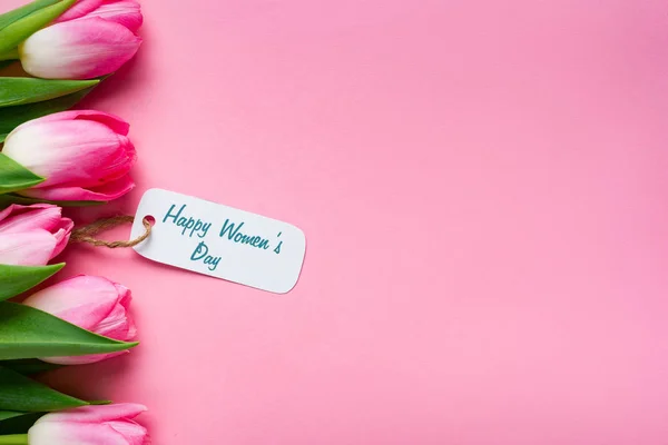 Top view of happy womens day lettering on paper label with row of tulips on pink background — Stock Photo