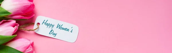 Top view of happy womens day lettering on paper label with tulips on pink surface, panoramic shot — Stock Photo