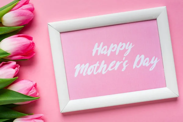 Top view of frame with happy mothers day lettering near tulips on pink background — Stock Photo