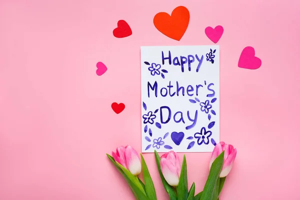 Top view of greeting card with happy mothers day lettering, tulips and paper hearts on pink background — Stock Photo