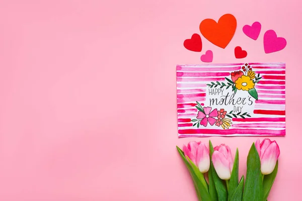 Vista superior de tulipanes, tarjetas de felicitación y corazones de papel sobre fondo rosa - foto de stock