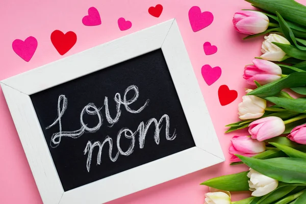 Вид сверху из картона с любовной мамой, лежащей рядом с бумажными сердечками и тюльпанами на розовом фоне — стоковое фото