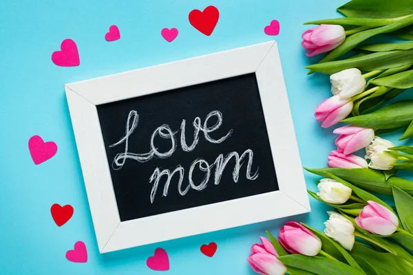 Vista superior de pizarra con letras de mamá del amor, corazones de papel y tulipanes sobre fondo azul — Stock Photo