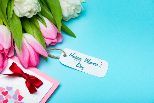 Bouquet of tulips with happy womens day lettering on paper label near greeting card on blue background — Stock Photo