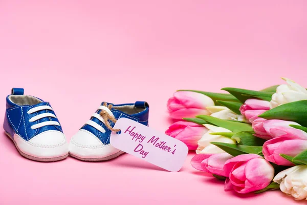 Baby booties with happy mothers day lettering on paper label near bouquet of tulips on pink surface — Stock Photo