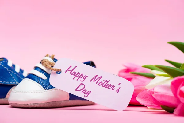 Nahaufnahme von Babybooties mit glücklichen Muttertag Schriftzug auf Papieretikett in der Nähe von Tulpen auf rosa Hintergrund — Stockfoto