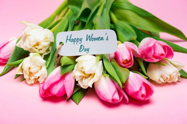 Lettering feliz día de las mujeres en la etiqueta de papel en el ramo de tulipanes sobre fondo rosa - foto de stock