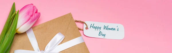 High angle view of envelope, paper label with happy womens day lettering and tulip on pink surface, panoramic shot — Stock Photo