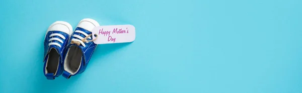 Top view of baby booties with happy mothers day lettering on paper label on blue surface, panoramic shot — Stock Photo