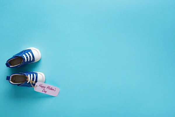 Vista dall'alto di etichetta di carta con felice giorno delle madri lettering e stivaletti bambino su sfondo blu — Foto stock