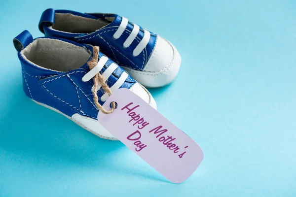 Visão de perto da etiqueta de papel com letras felizes do dia das mães e botas de bebê na superfície azul — Fotografia de Stock