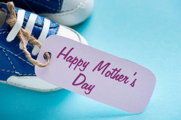 Vista de cerca de las madres felices letras del día en la etiqueta de papel y botines de bebé en la superficie azul - foto de stock