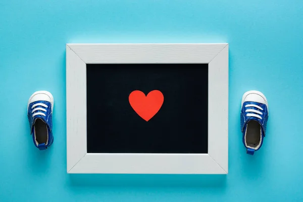 Vue du dessus des chaussons bébé avec coeur en papier sur tableau à la craie sur fond bleu, concept de fête des mères — Photo de stock