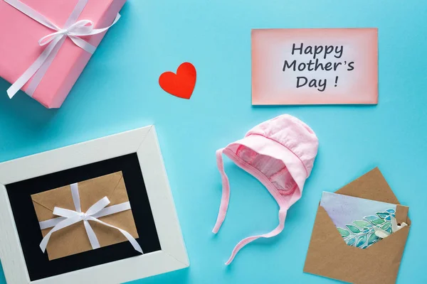 Vista superior de la caja de regalo, sobres y tarjetas de felicitación con letras felices del día de las madres y sombrero del bebé sobre fondo azul - foto de stock